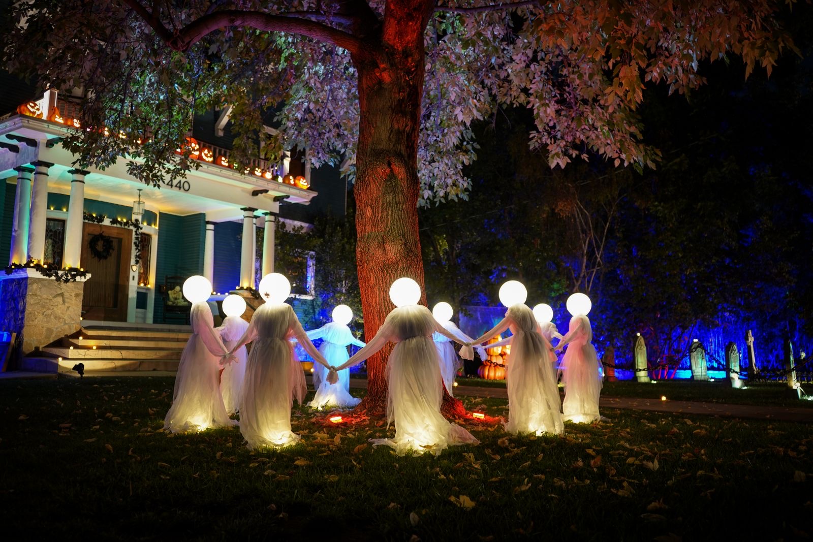 Glowing ghost decorations circle a tree in front of a house lit up for Halloween at night