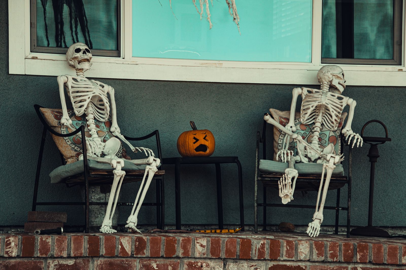 Two skeleton decorations lounging in porch chairs beside a carved pumpkin