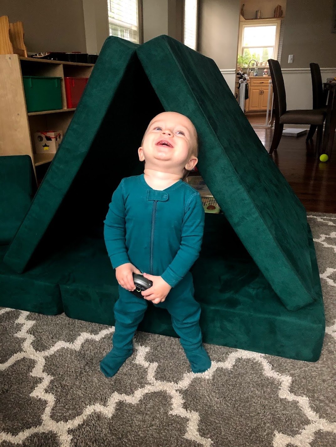 Toddler standing inside a green Nugget play couch built into an A-frame tent fort on a playroom rug